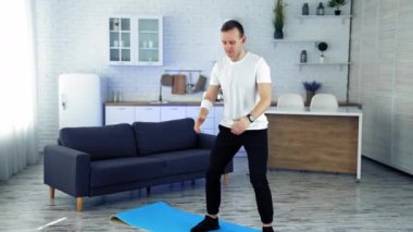 Ev antrenmanı. Beyaz tişörtlü ve siyah pantolonlu genç adam mutfak içinde çömelme egzersizi yapıyor. Spor eğitimi kapalı alanlarda. Yavaş çekim.