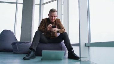 Pencere arkasında aletleri olan genç bir adam. Yakışıklı adam ofisteki koltukta oturuyor ve cep telefonu kullanıyor. Dizüstü bilgisayar yerde. Serbest konsept.