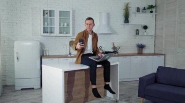 Evde online çalışan genç bir adam. Mutfak masasında dizüstü bilgisayarla oturan ve telefonla video görüşmesi yapan serbest çalışan bir erkek. Uzak görev.