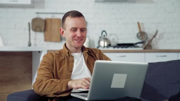 キッチンでラップトップを使ったフリーランサー ラップトップを介して会話をする幸せな男 自宅で働くオンライン コロナウイルス検疫中の自宅からの遠隔作業 — ストック動画