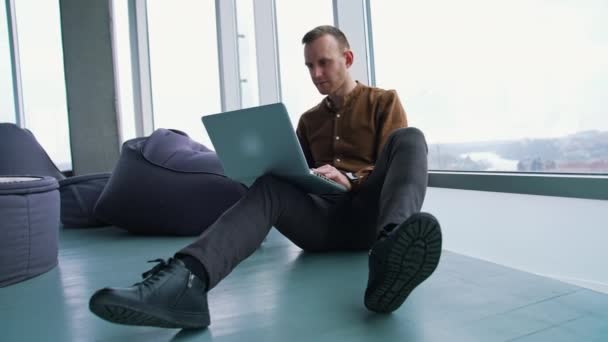 ラップトップで働いている自信のある男性 若いビジネスマンが床に座って 昼間の窓の近くのワイヤレスコンピュータにタイピングします 真剣なフリーランサーは屋内で働く — ストック動画