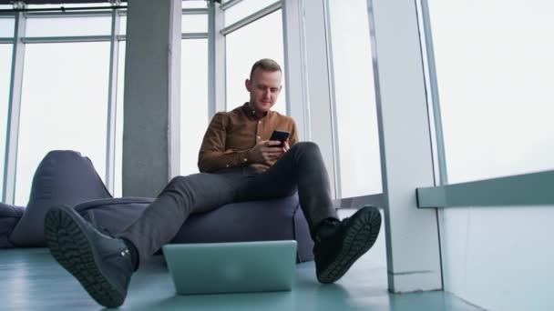 若者は携帯電話を使っている ビジネスマンは大きな窓の背景に携帯電話を持ってオットマンに座っています オフィスの床にラップトップ 下からの眺め — ストック動画