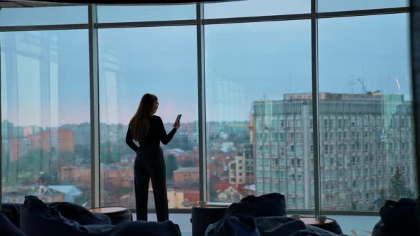 携帯を持った若いビジネスマン 大きな窓の近くに近代的なオフィスの女性のリアビュー 街の背景を持つパノラマウィンドウビュー バックビュー — ストック動画