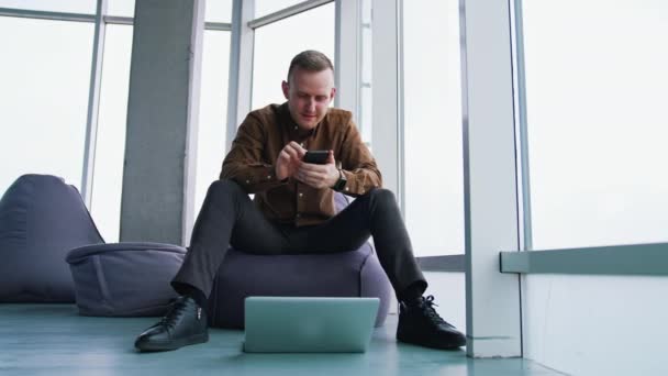 窓の背景にガジェットを持っている若い男 ハンサムな男がオットマンに座って 携帯電話を使っている 床の上のラップトップ フリーランスコンセプト — ストック動画