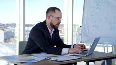 Dizüstü bilgisayar kullanan ciddi bir erkek işçi. Büyük pencereli bir ofiste dizüstü bilgisayarla çalışan odaklanmış bir iş adamı. Bilgisayarda daktilo eden gözlüklü bir iş adamı..