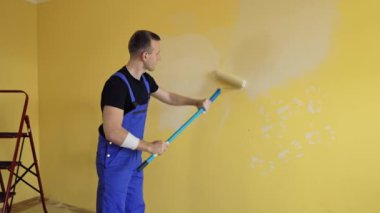 Adam evdeki duvarları boyuyor. Mavi üniformalı genç inşaatçı uzun saplı boya silindirli duvarları yeniliyor. Resim hizmetleri.