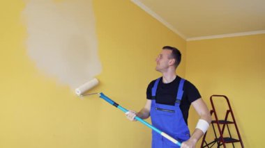Yeni bir renkle oda geliştirme. Profesyonel işçi mavi tulumlu, uzun saplı boya silindirli duvarı boyuyor. Çalışan bir zanaatkar.