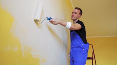Boyacı duvarları silindirle boyuyor. Tulumlu genç bir işçinin portresi. Odada yeni bir renkle makyaj yapıyor. Profesyonel resim hizmetleri.