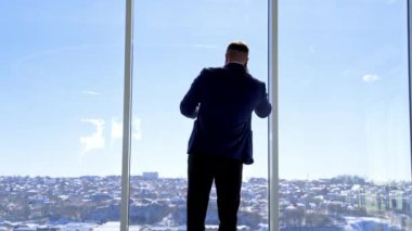 İş adamı telefonda sohbet ediyormuş. Zarif takım elbiseli genç adam büyük ofis penceresinin yanında duruyor ve telefonla konuşuyor. Arka görünüm.