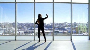 Kız ofiste selfie çekiyor. Güneşli bir günde büyük bir ofis penceresinin yanında dururken cep telefonuyla fotoğraf çeken genç bir kadın. Arka görünüm.