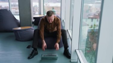 Pencerenin yanında dizüstü bilgisayarı olan genç bir adam. Tişörtlü yakışıklı adam rahat bir koltukta oturuyor ve dizüstü bilgisayarına bakıyor..