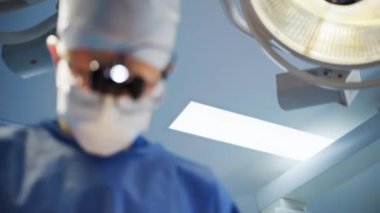 Ameliyat sırasında cerrahın yüzü. Maskeli profesyonel bir doktor ve bulanık geçmişi olan bir hastaya ameliyat yapan cerrahi ışıklar. Yakın plan..