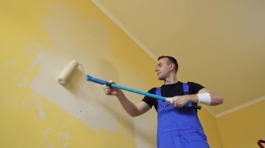 İşyerindeki ressam bir silindirle duvar boyuyor. Mavi tulumlu bir işçi bir odayı yeniliyor. Profesyonel boyama hizmeti sunuyor. Aşağıdan görüntüle.