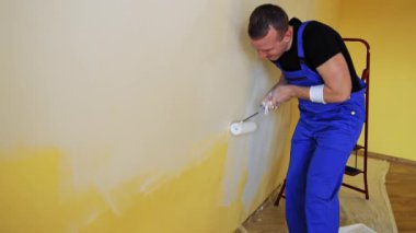 Genç adam bir silindirle beyaz bir duvar boyuyor. İçerisinin tamiri. Erkek dekoratör boş odada bir duvar boyuyor. Tulumlu bir ressam odayı yeniliyor..