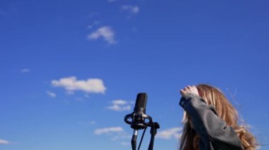 Güzel bir kadın dışarıda tek başına şarkı söylüyor. Mavi gökyüzü arka planında mikrofonu olan genç şarkıcı kız. Aşağıdan görüntüle.