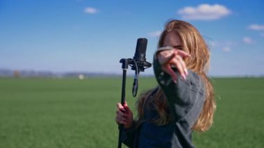 Yeşil tarlada güzel bir şarkıcı kız. Çekici sarışın kadın mikrofona şarkı söylüyor ve kameraya bakıyor..