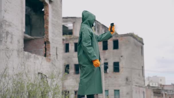 携帯電話付きの保護スーツとガスマスクで人間 放射線の間に破壊された場所でセルフィーをする安全制服 手袋および呼吸器の認識できない人 — ストック動画