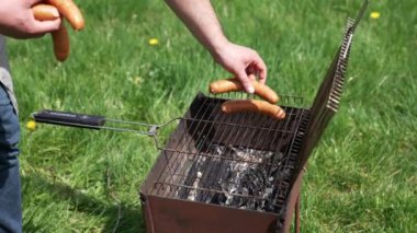 Adam kızartma için ızgaraya sosis koyar. Sosisli sandviçi ateşte pişirmek. Doğada barbekü. Yeşil çim arka planda Brazier.