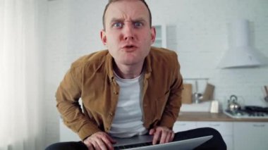 Evde çalışan komik komedyen. Mutfak masasında dizüstü bilgisayarla oturmuş kızgın bir şekilde kameraya bakan genç bir serbest çalışanın kızgın yüzü..