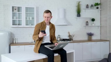 Evde çalışan serbest çalışan bir adam. Mutfak masasında dizüstü bilgisayarla oturan ve cep telefonuyla fotoğraf çeken genç bir adam..