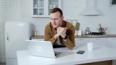 Genç adam mutfak arka planında görüntülü konuşma yapıyor. Freelancer evdeki kablosuz bilgisayarda çalışıyor. Uzak iş.
