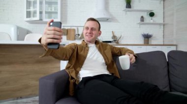 Genç adam evde kendi fotoğrafını çekiyor. Kanepede oturan yakışıklı adam cep telefonu kullanıyor ve içeride çay içiyor. Gülümseyen adam karantina sırasında evde oturmaktan zevk alıyor..