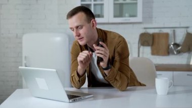 İnternetten alışveriş. Genç adam dizüstü bilgisayarın önünde konuşuyor ve yeni bir cihaz öneriyor. Evdeki dizüstü bilgisayarda çalışan gülümseyen adam. Uzak görev.