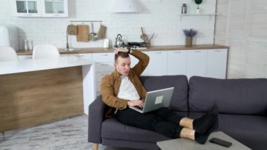 Genç adam evde bilgisayar oyunu oynuyor. Mutfak arka planında dizüstü bilgisayarla kanepede yatan gergin adam..