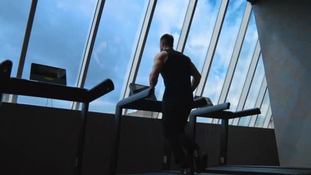 空の背景にあるトレッドミル屋のスポーツマン フィットネス男はジムでシミュレーターで走り 窓から眺めている リアビュー — ストック動画