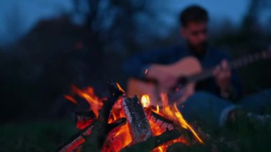 Yanan şenlik ateşi. Genç adam karanlıkta ateşin yanında gitar çalıyor. Pikniğin rahat atmosferi ateşin yanında..