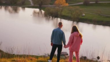 Genç bir adam ve kadın batan güneşe bakıyor. Gün batımında nehrin üzerindeki tepede el ele tutuşan romantik çift. Arka görünüm.