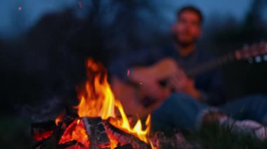 Akşam şenlik ateşinde. Geceleri kırmızı ve turuncu alevler. Gitar çalan bir adamın bulanık arka planında parlak kamp ateşi.