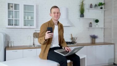 Bilgisayarı ve telefonu olan bir öğrencinin portresi. Gülümseyen genç adam mutfak masasında oturuyor ve evde dizüstü bilgisayar ve cep telefonu kullanıyor. Karantina konsepti.