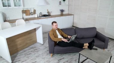 Evdeki kablosuz bilgisayarda çalışan rahat bir adam. Genç bir adam kanepeye uzanıyor ve modern mutfak arka planında dizüstü bilgisayar kullanıyor. Karantinada iş.