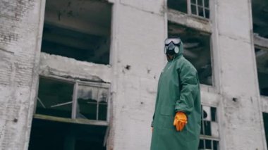 Koruyucu giysili ve gaz maskeli bir adam yıkılmış binanın arka planında duruyor. Kıyametten sonraki dünya kavramı..