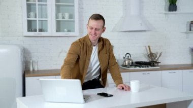Genç adam mutfakta çalışır ve çay içer. Karantina sırasında evde ders çalışan mutlu bir öğrenci. Adam rahat bir dairede iş için dizüstü bilgisayar kullanıyor..