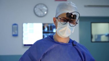 Gözlüklü bir doktorun portresi. Dürbünlü büyüteç takan ve ameliyathanede kamerada konuşan bir cerrah..