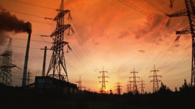Günbatımında yüksek voltaj kablosu ve pilonlar. Kırmızı batan güneşli dramatik gökyüzüne karşı elektrik kulelerinin siluetleri.
