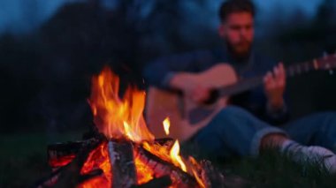 Geceleri çok güzel kamp ateşi. Akşamları gitar çalan bir adamın arka planında kırmızı ve turuncu alevler yanıyor. Dışarıda boş vakit geçirmek..