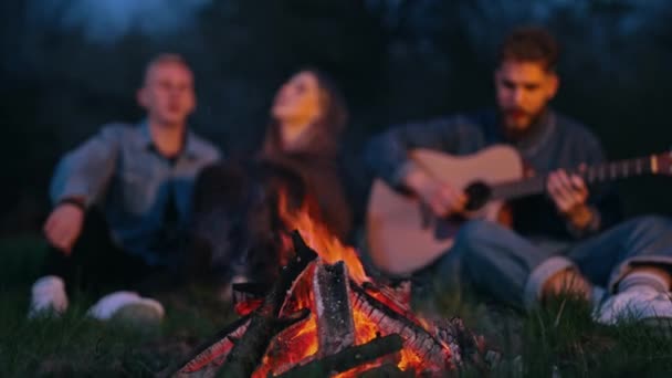 火の近くの友人の居心地の良い夜 夜にギターに歌う若者のぼやけた背景に暖かい赤いキャンプファイヤー — ストック動画