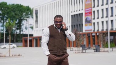 Neşeli iş adamı dışarıda telefonla konuşuyor. Gözlüklü, beyaz gömlekli ve kahverengi kostümlü Afro-Amerikan bir adam şehirde yürüyor ve telefonla konuşuyor..