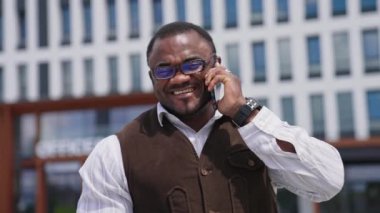 Pozitif Amerikan erkeklerinin telefonlu portresi. Şık gözlüklü Afrikalı Amerikalı işadamı telefonda konuşuyor ve şehirde kameraya gülümsüyor..