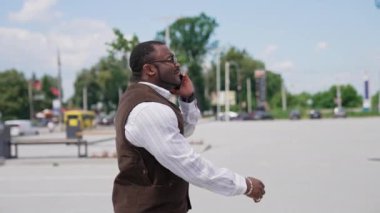 Zenci işadamı açık havada iş başında. Şehirde yürüyen ve telefonla konuşan Afro-Amerikan tarzı bir adam. Profil görünümü.