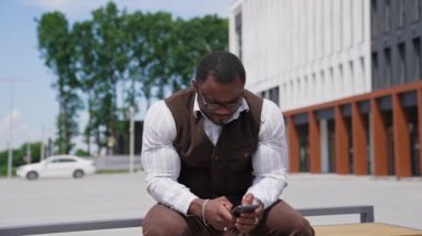 Telefona bakan ciddi siyah derili adam. Takım elbiseli, açık havada oturan ve cep telefonu kullanan Afrikalı Amerikalı adamın portresi..