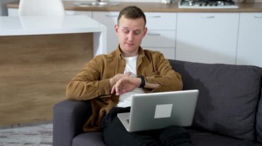 Kanepede yatarken dizüstü bilgisayar kullanan serbest çalışan bir erkek. Genç adam saatine bakıyor ve evde bir dizüstü bilgisayar üzerinde çalışırken biriyle konuşuyor..