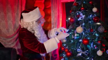 Noel Baba Noel ağacını süslüyor. Noel Baba kostümü giyen beyaz sakallı yaşlı adam Noel balolarını süslenmiş yılbaşı ağacında düzeltiyor. Konforlu evde Noel atmosferi.