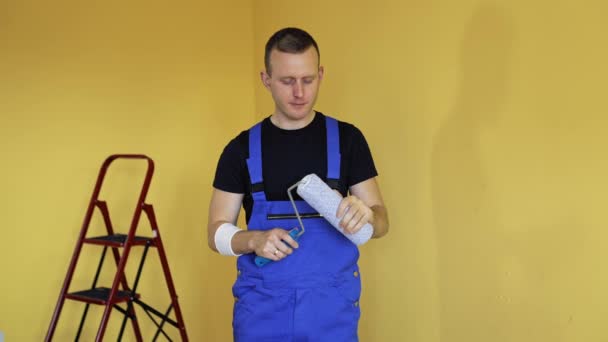 一个工人画家的肖像 身穿蓝色工作服的年轻人站在房间里 拿着滚筒刷子 然后在公寓里化妆 住房改善和翻新 — 图库视频影像