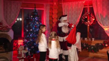 Noel Baba ve çocuklar geldi. Beyaz sakallı mutlu Noel Baba koltuğa oturur ve çocukları dinler. Noel dekore edilmiş oda arka planı.