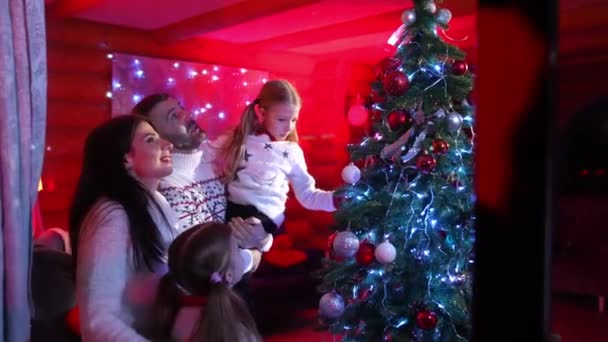 幸せな家族がクリスマスを祝います 親とその子供たちは 光と明るいボールで飾られたクリスマスツリーの近くで一緒に時間を過ごします ウィンターホリデー — ストック動画