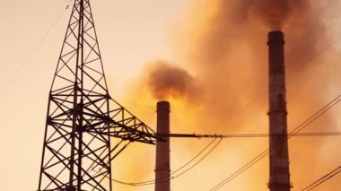 Fabrika borularından çıkan turuncu dumanlar. Kalın bacalardan gelen kirli emisyon bulutları havayı dolduruyor. Endüstrinin kara dumanının arka planında elektrik kulesi.